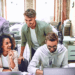 A group of young professionals collaborating and smiling around a laptop in a modern open office environment.