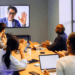 A diverse team in a modern office conference room attends a virtual meeting on a large screen, collaborating with a remote colleague via video call.