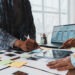 Two UX designers collaborate on mobile app wireframes using printed layouts, sticky notes, and a tablet, with interface mockups displayed on a laptop.